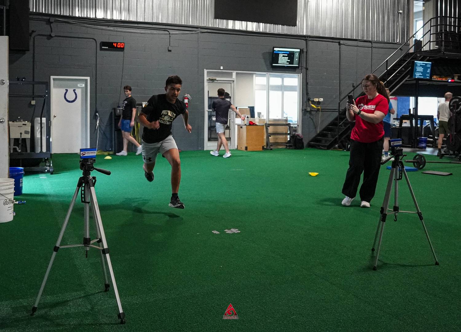 A high school athlete sprinting between electronic timing gates on the turf at Athletes Acceleration while a coach records the run